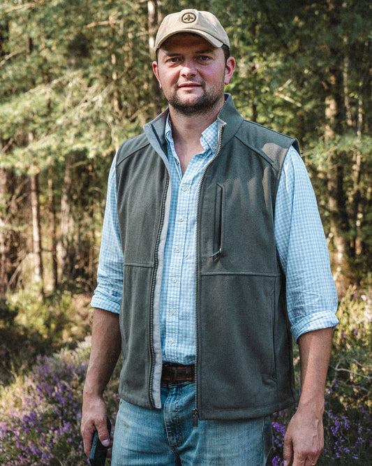 Torr Wool Gilet in Heather Green with a Rollock Canvas Cap in Tan, worn open over a pale blue check shirt among the pines.