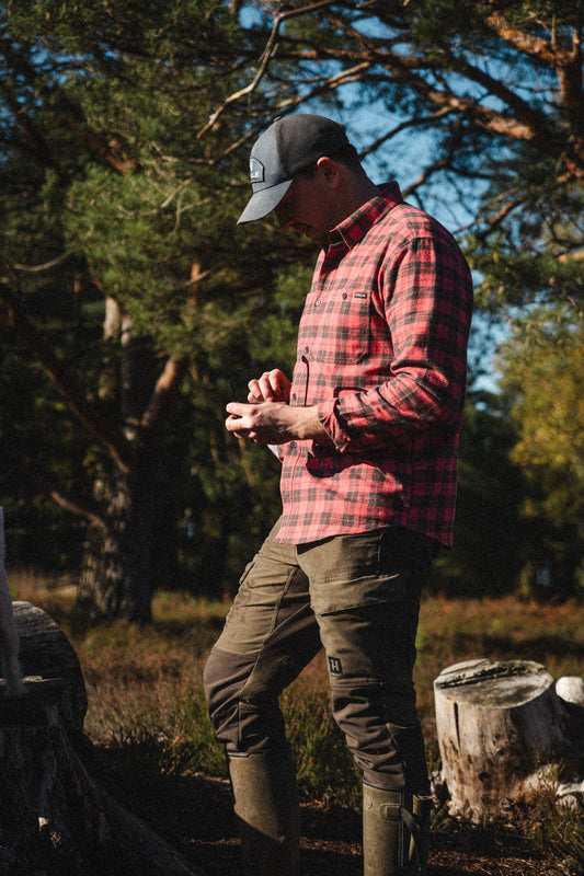 A man in a red check Brae Flannel Shirt and a grey trucker cap, standing in the sun among pine trees.