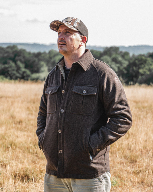 Man in the Arkle Wool Jacket and a camo trucker cap, hands in pockets, looking out across a field of dry grass towards distant woodland.
