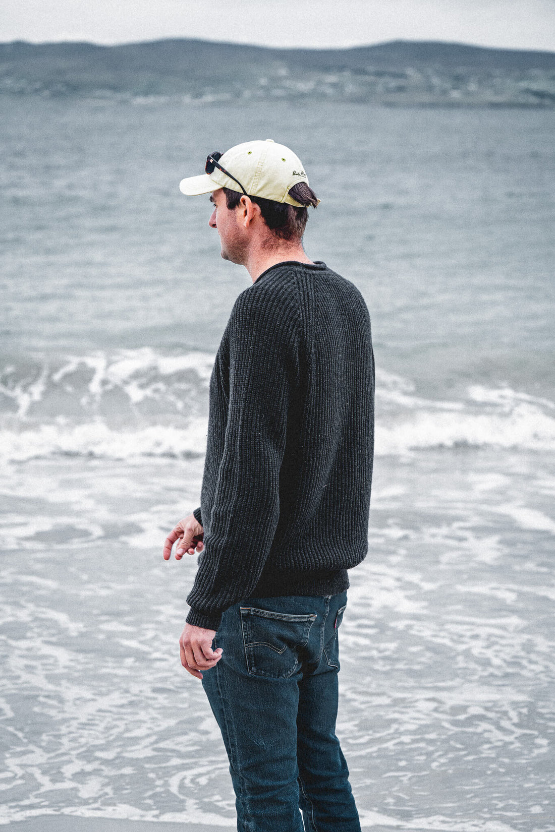 Man in the Tarbert Fisherman's Jumper in Slate and a pale cap standing at the water's edge, looking out over breaking waves towards low hills.
