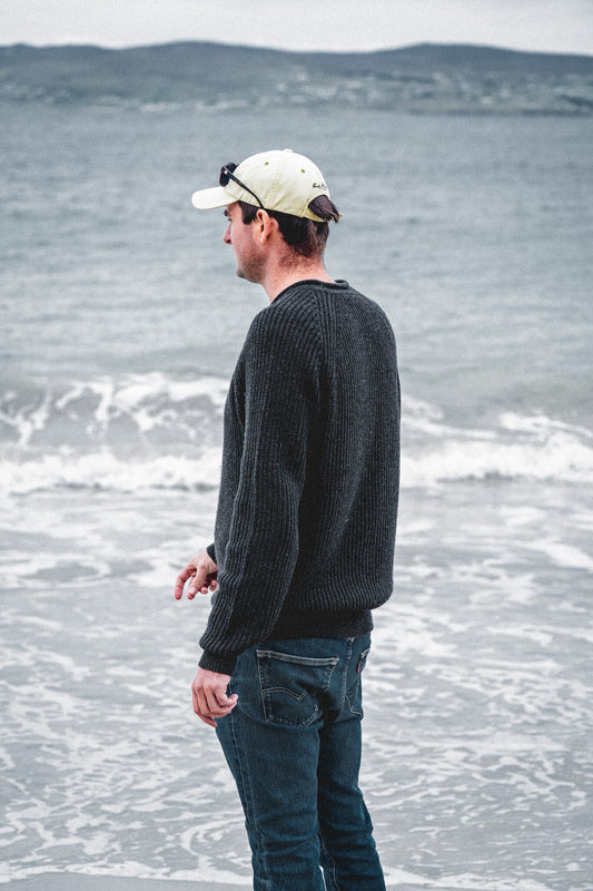 Man in the Tarbert Fisherman's Jumper in Slate and a pale cap standing at the water's edge, looking out over breaking waves towards low hills.