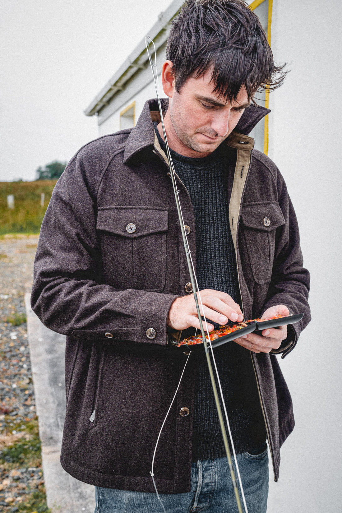 Arkle Wool Jacket, worn over a navy knit while choosing a fly from an open fly box, rod in hand.