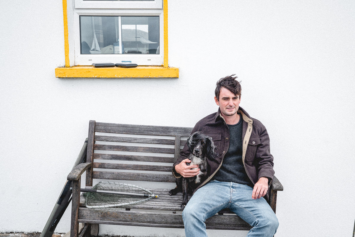 Arkle Wool Jacket, worn sitting on a weathered bench with a spaniel, a landing net and rod resting alongside.