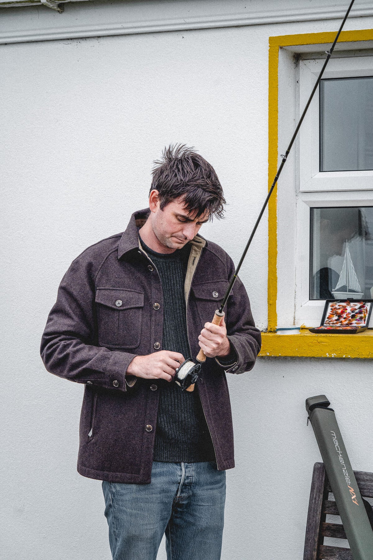 Arkle Wool Jacket over a Tarbert Fisherman's Jumper in Slate, worn fitting a reel to a fly rod outside a whitewashed cottage.